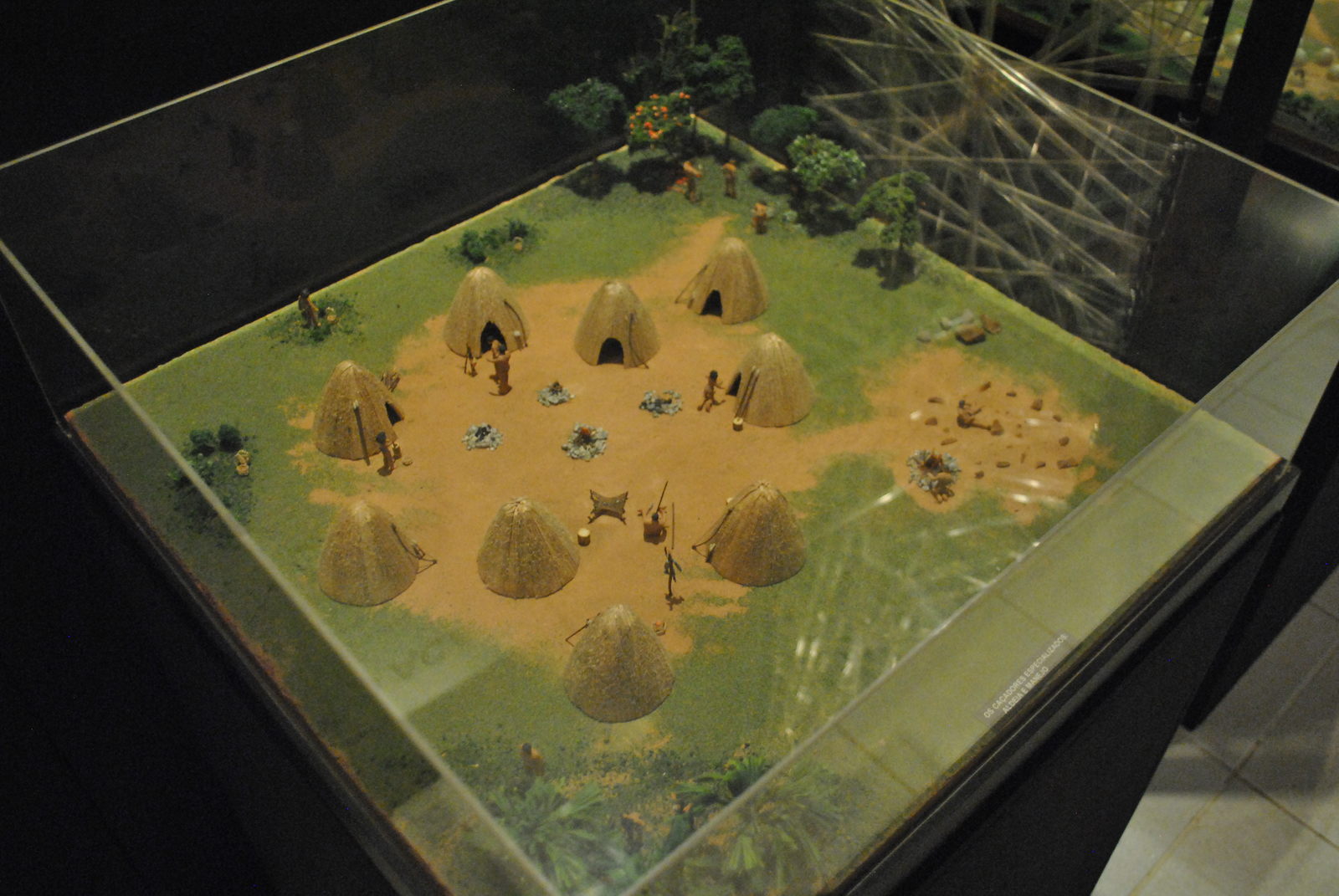 Foto de maquete ilustrando como eram as atividades dos povos antigos, com as casas organizadas em circulos, ao centro fogueiras acesas e índios trabalhando em
												 diversas atividades, como cozer alimemntos, manutenção da esturas da casa, cortes de lenhas entre outras atividades